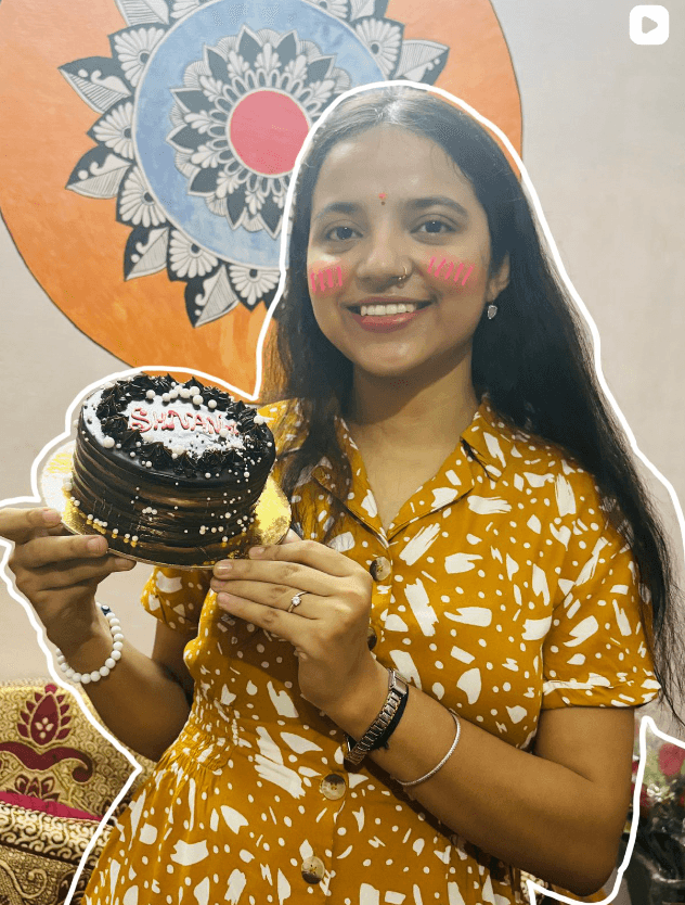 Shivangi holding a small chocolate cake with SHIVANGI written in icing, mandala art behind her