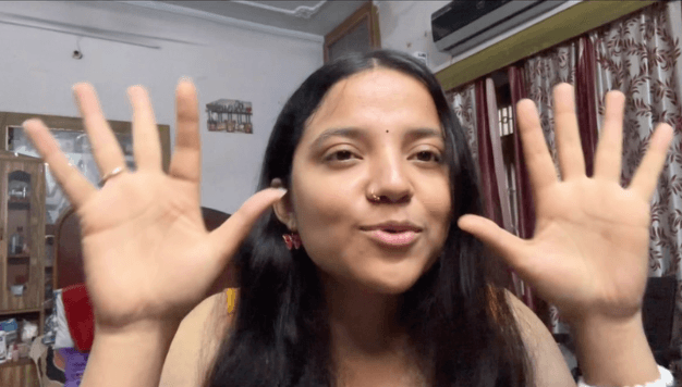 Shivangi in a yellow top holding both hands up with fingers spread and butterfly earrings, smiling at the camera